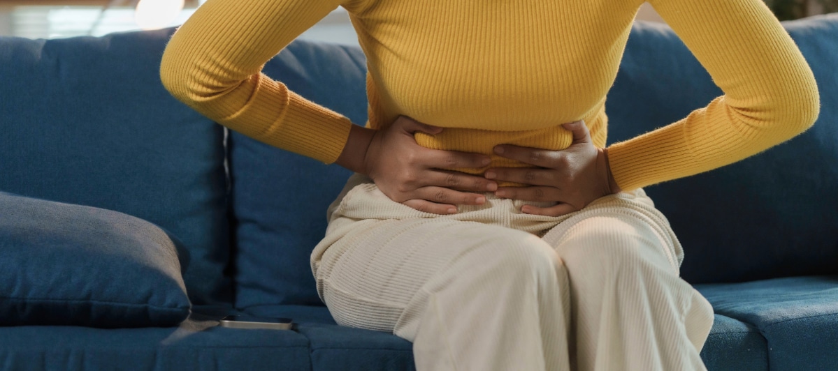 A Guide to Chronic Pelvic Pain Causes and Treatments-3-Pain A woman sits on a couch and holds her midsection due to chronic pelvic pain