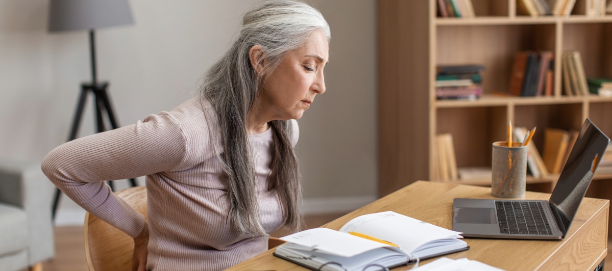 Effective Pain Relief Strategies for Spinal Stenosis-3-Back Pain A woman sitting at a table holds her back in pain from spinal stenosis