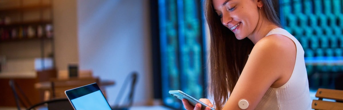 Four Benefits of a Diabetes Management Plan-3 A woman smiles as she checks her glucose levels while sitting at a desk as part of her diabetes management plan