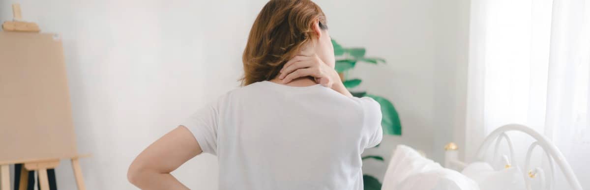 Heart Disease Symptoms You Need to Know (18) A woman sits on her bed and holds her neck due to upper middle back pain