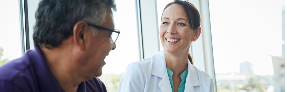 Heart Disease Symptoms You Need to Know (2) A doctor in a lab coat smiles as she talks to her patient about heart disease risk factors