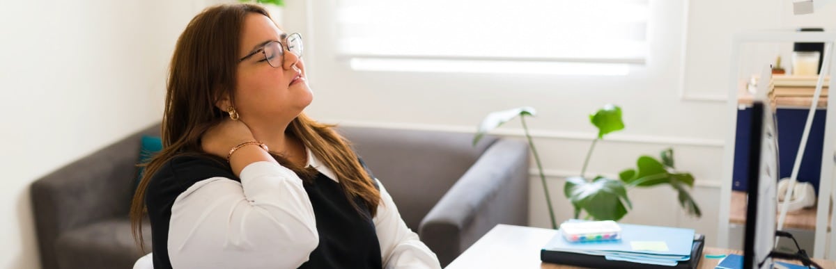 Heart Disease Symptoms You Need to Know (22) A woman sitting at an office desk holds her neck due to upper middle back pain