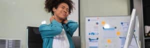 A woman at an office desk winces and holds her neck due to upper back pain