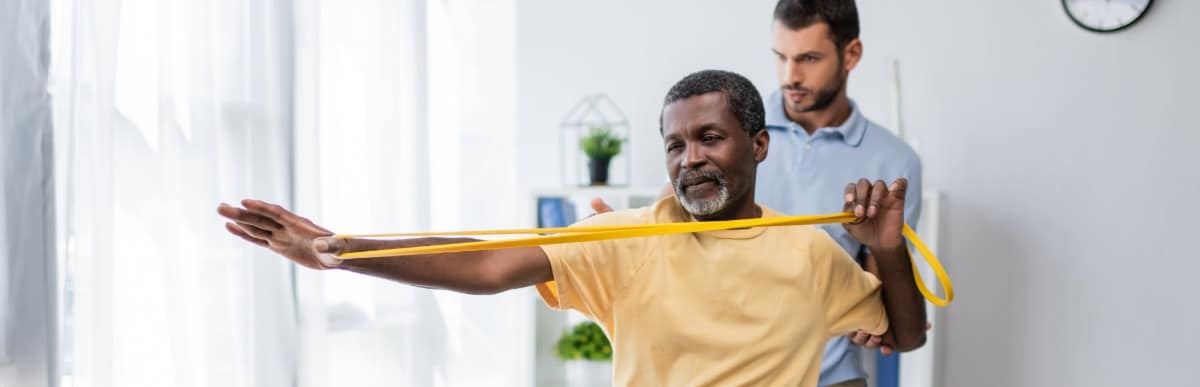 Heart Disease Symptoms You Need to Know (28) A man stretches his shoulder with a a strap under the supervision of a physical therapist