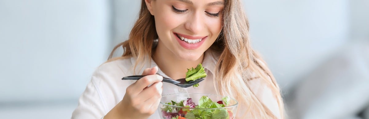 Heart Disease Symptoms You Need to Know (3) A woman smiles as she takes a forkful of a salad to help avoid heart disease risk factors