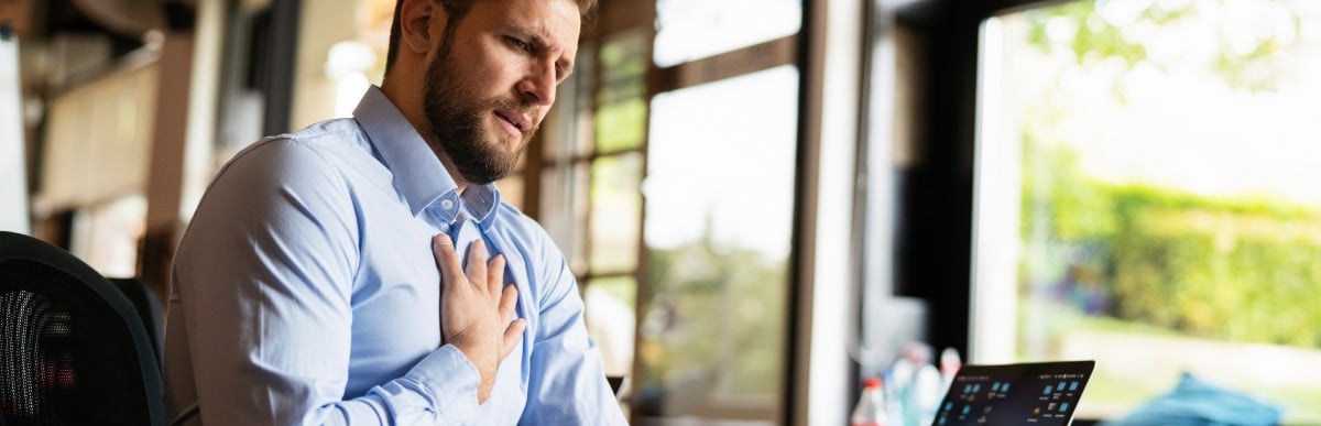 Heart Disease Symptoms You Need to Know-3 A man sitting at a desk holds his chest due to chronic pain, which is one of the heart disease symptoms