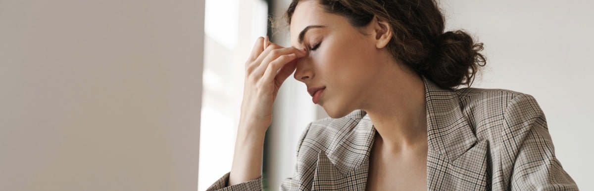 Heart Disease Symptoms You Need to Know (51) A woman sitting at an office desk holds her nose and closes her eyes due to fatigue which is one of the early signs of autoimmune arthritis