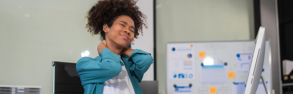 Heart Disease Symptoms You Need to Know (68) A woman sitting at an office desk and holds the back of her neck in pain