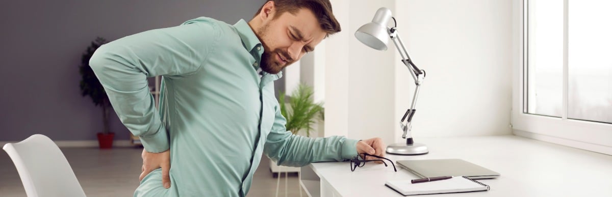 Heart Disease Symptoms You Need to Know (72) A man sitting at a desk winces in pain and holds his back as he searces online for vertebral compression fracture treatments