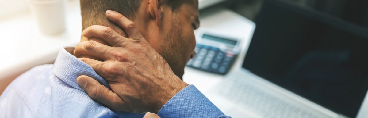 Heart Disease Symptoms You Need to Know (84) A man sitting at an office desk rubs his neck in pain due to myofascial pain syndrome