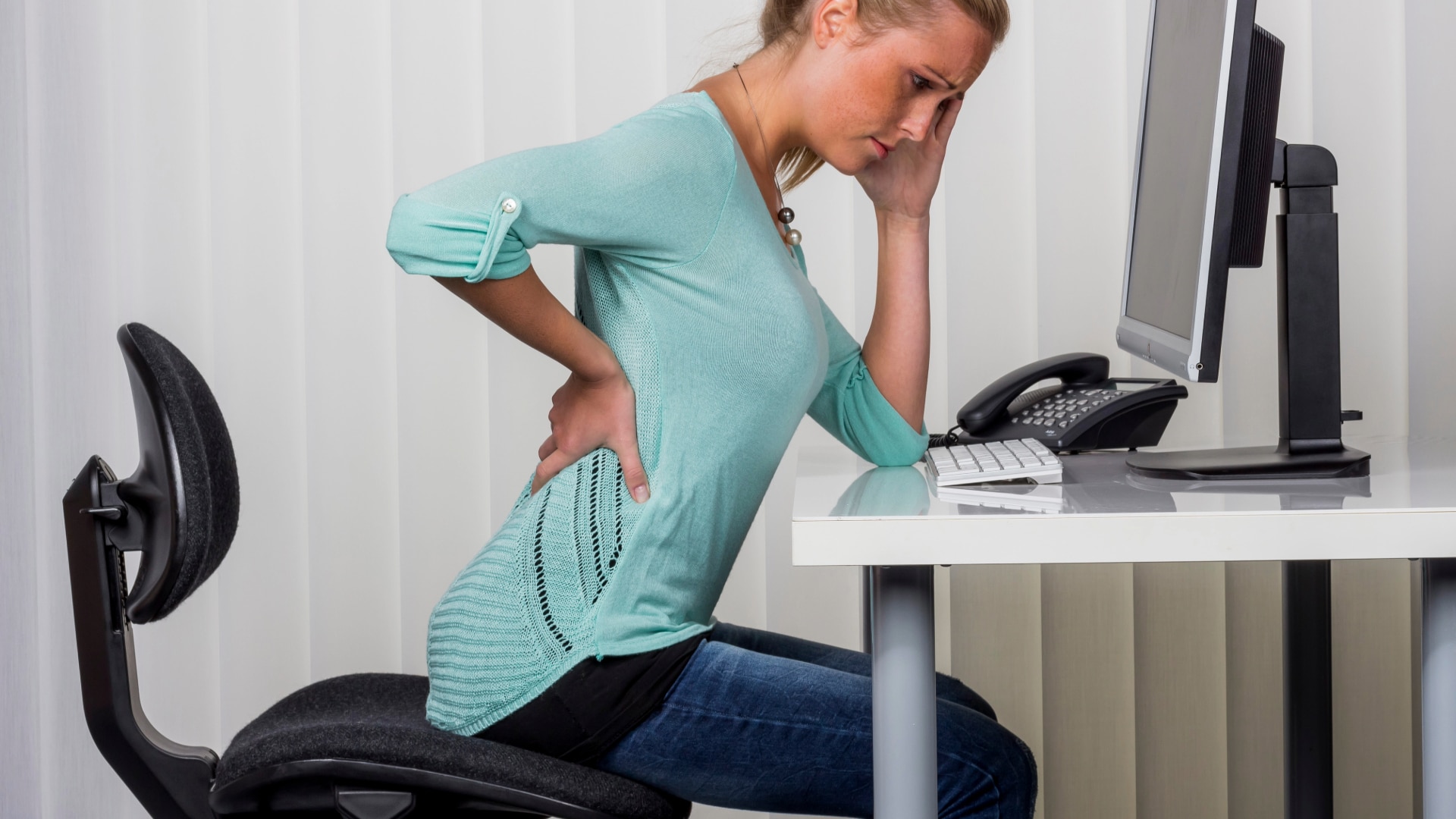 Hip Bracing-2 A women in a green shirt sitting at her desk and holding her lower back while suffering hip pain