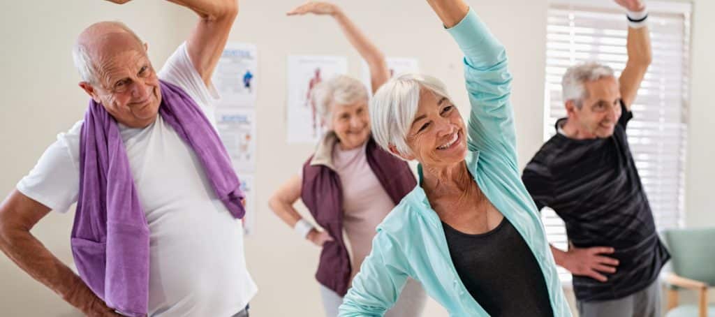 A group of older people smile as the stretch their hands in their air in an exercise class