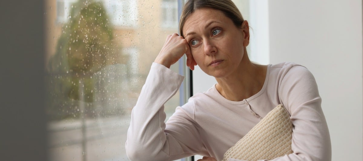 The Relationship Between Inflammation and Weight Gain-3-Sad A woman puts her hand to her head and looks out a window as she lives with inflammation and weight gain