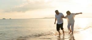 An older couple holds hands and smile as they walk barefoot on a beach