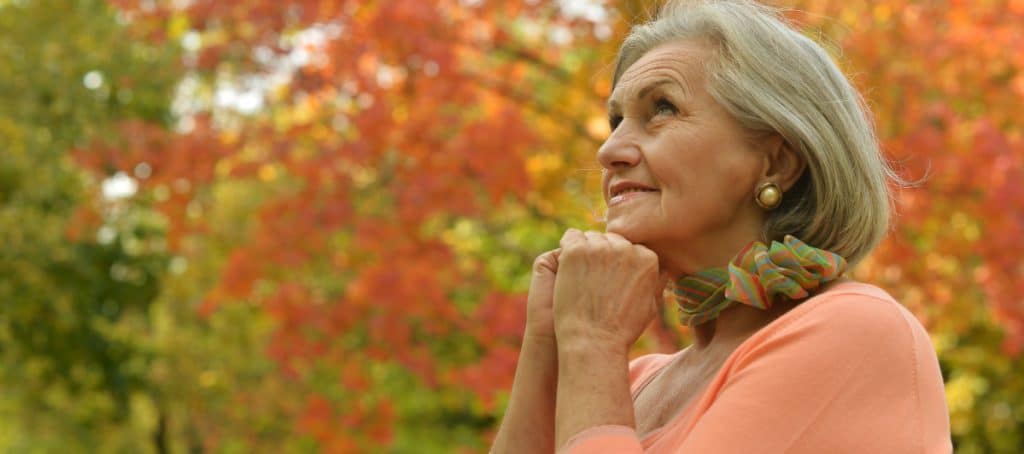 A woman smiles with her hands under her chin while standing in a forest
