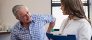 A man sitting on an exam table grabs his back in pain as he talks to the doctor about kyphoplasty